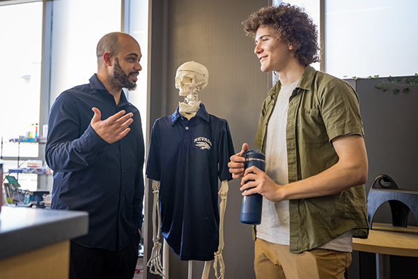 A professor and a student talk in a lab setting next to a skeleton model dressed in a university shirt. 
