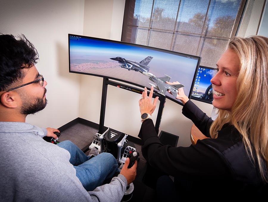 A student sits at a flight simulator, looking at a monitor with a plane on the screen and his right hand holding a control while an instructor sits next to him, holding a model plane and pointing to it.