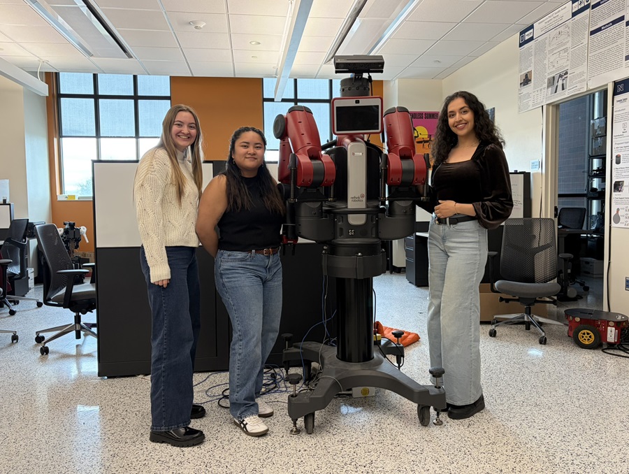 Three women stand in a lab with a tall robot on wheels.