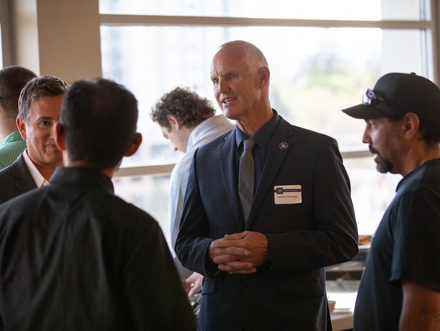 A man in a suit with a nametag reading "Gordon Boettger" stands among three men, talking; in the background two people facing away from the camera look down at a buffet table.
