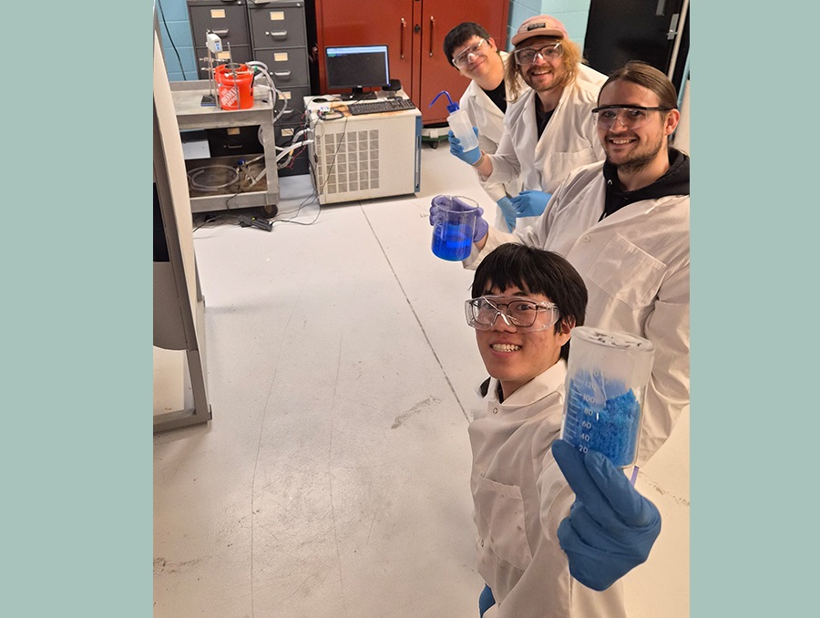 Four students in lab coats, gloves and protective goggles stand in a lab; the one in the front holds a container with blue crystals, the second holds a cylinder of blue liquid.