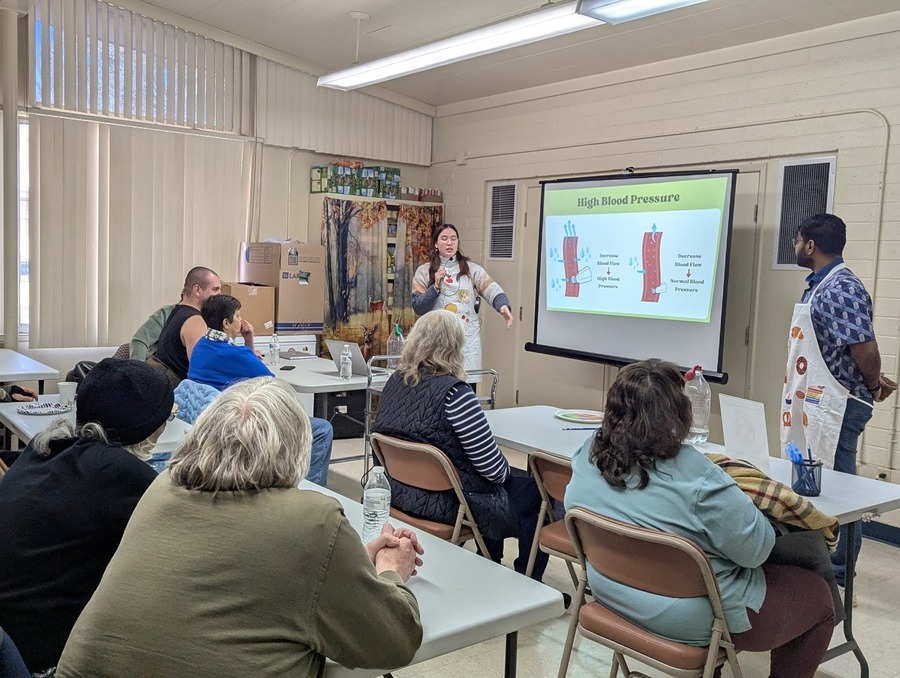 A man and a woman stand on either side of a presentation screen while speaking to an audience.