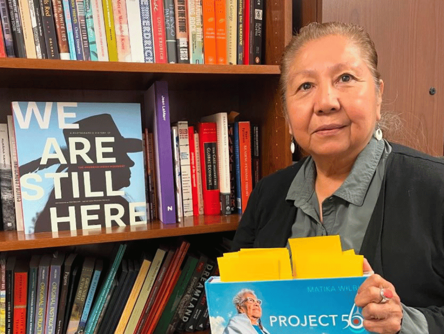 Debra Harry holding a book, next to a bookshelf and book titled "We Are Still Here.". 