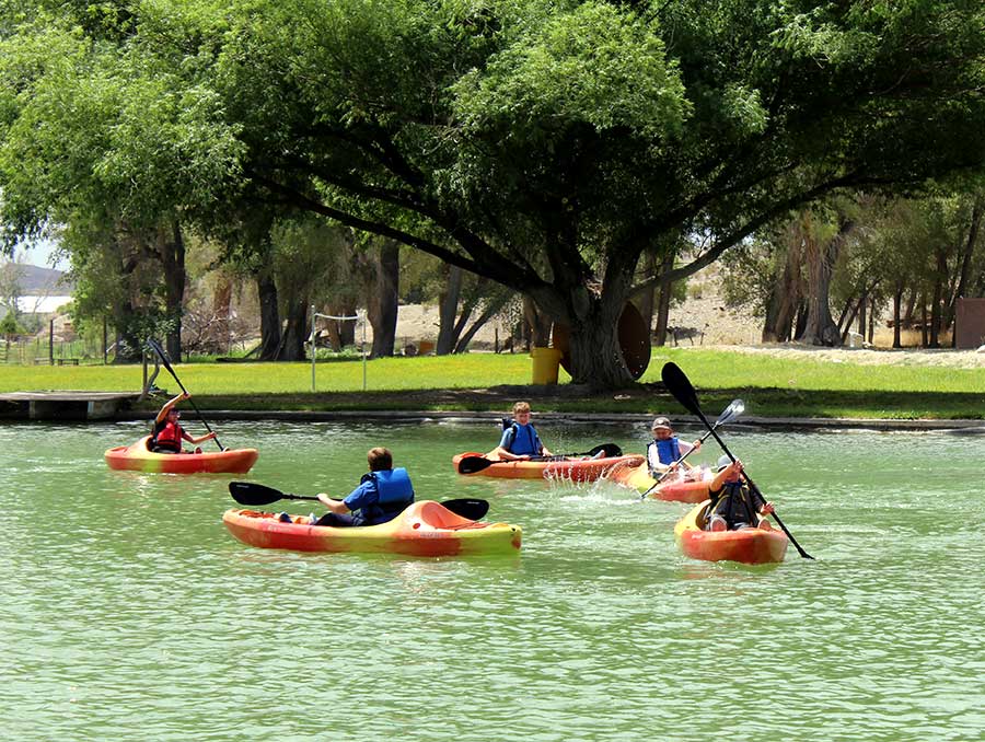 In a pond, surrounded by green grass and large shade trees, five youth in lifejackets, baseball caps and sun shirts splash in the water as they paddle their own yellow and orange sit-on-top kayaks.