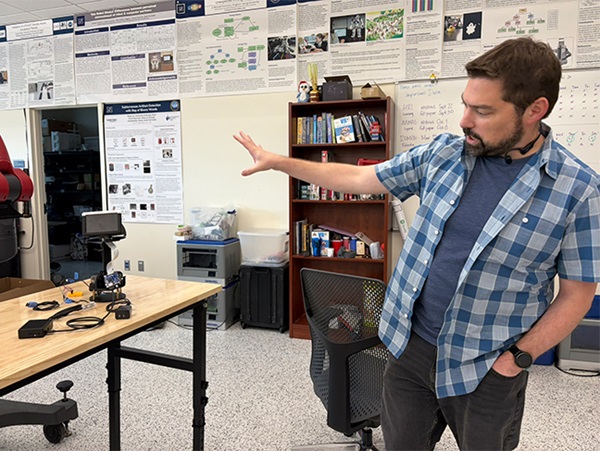 A man standing in a lab gestures to a table bearing technical equipment while talking. 