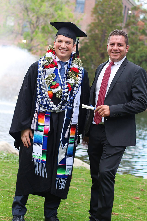 Retana wearing a cap, gown and multiple honor cords stands outdoors next to another man in a suit holding a diploma on commencement day.