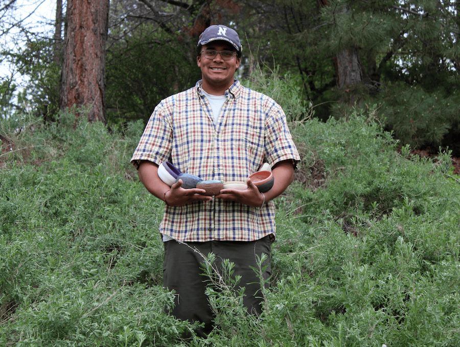 Grayson stands outside in a forest holding a set of six ceramic pots he made.