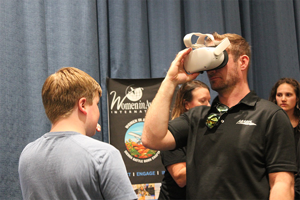 A man holds a virtual reality headset up to his eyes while a boy looks on; behind them is a banner reading "Women in Aviation" and two women standing.