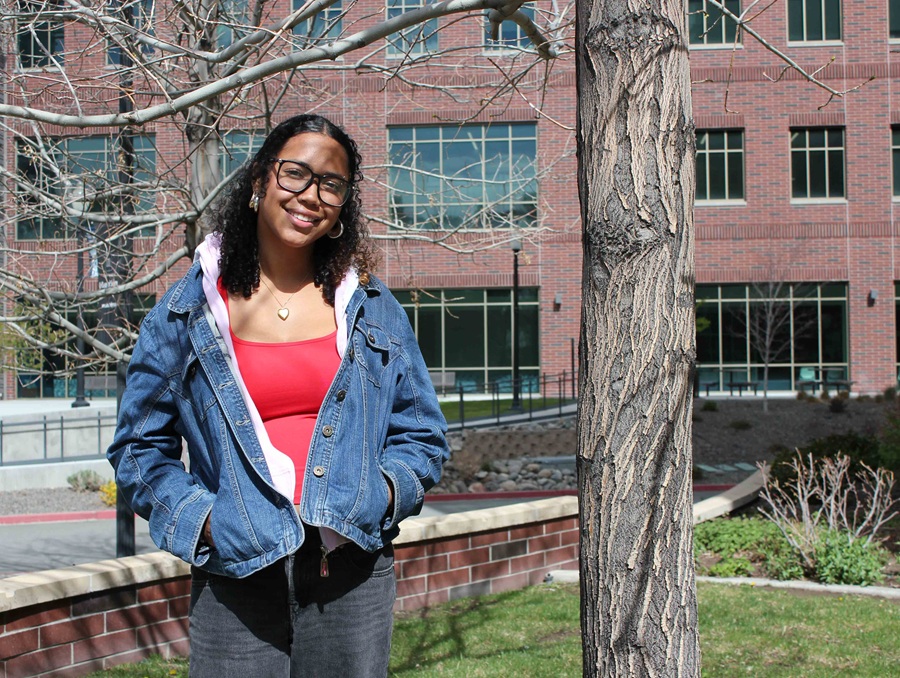 A young woman in a red top and blue jacket poses in front of the William Pennington Engineering Building, a tree to her left.