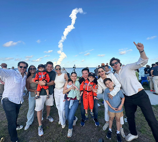 A group of adults and children smile and pose outdoors near the ocean as a rocket launches in the background, leaving a trail of smoke in the sky.