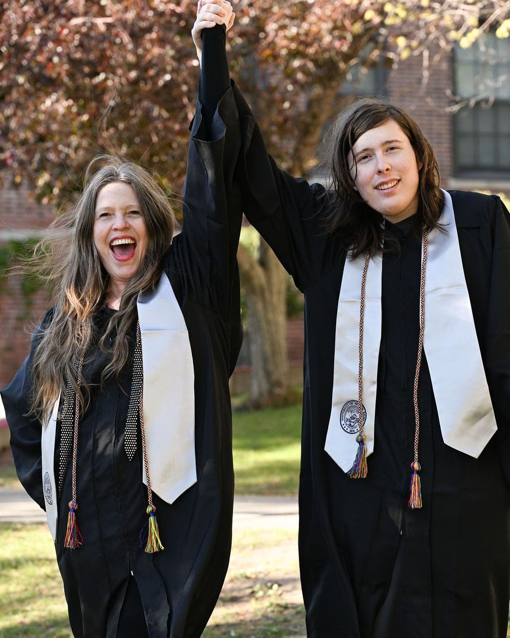 Mother and son, wearing graduation gowns, holding hands.