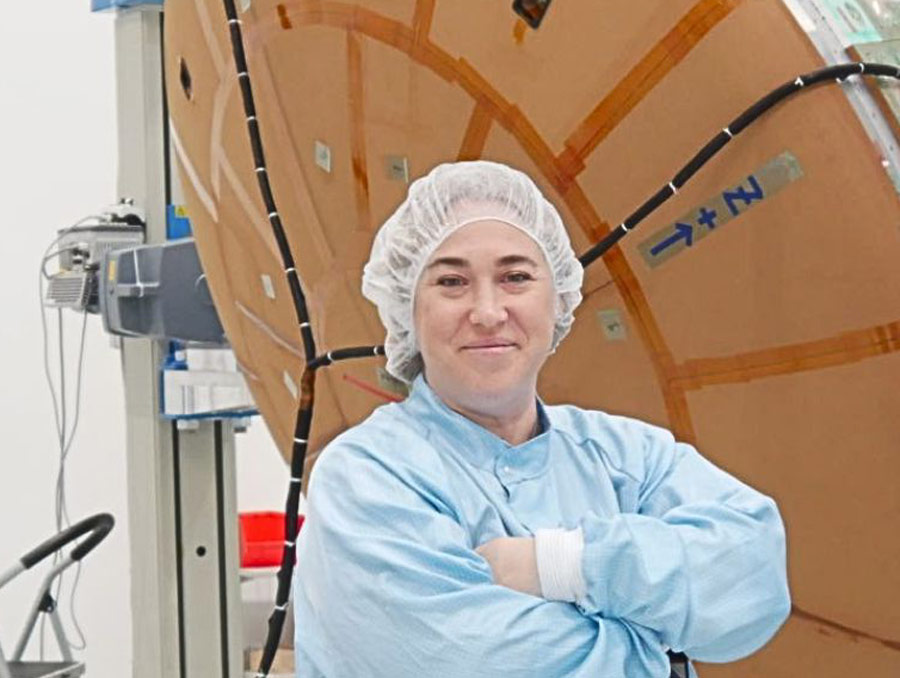 A person wearing a clean suit stands with their arms folded, smiling for the camera, in front of a large orange disk.