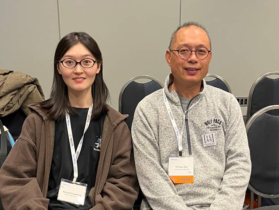 Xiang Xiang Wang and Tin Yau Tam wearing conference lanyards and sit in a row of chairs, smiling for the photo.