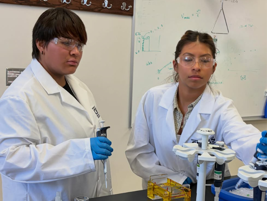 Two students wearing lab coats operate lab equipment in a classroom.