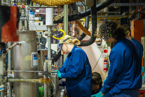 Scientists wearing gloves, safety goggles and blue lab coats work on equipment in a crowded room.