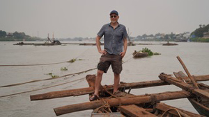 A man stands on the bow of a wooden boat on a river.