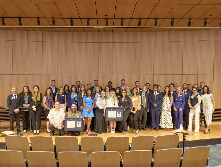 Group photo of 44 University of Nevada, Reno Online Executive MBA graduates standing on a stage in the John Tulloch Business Building following the program's 2026 graduation ceremony. Several graduates in the front row hold framed diplomas.