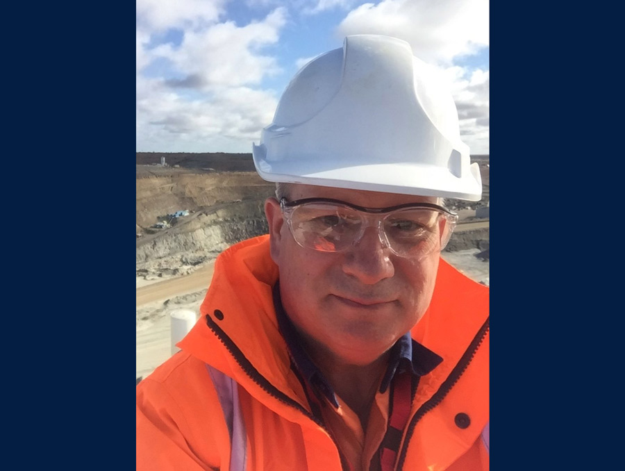 A man wearing a white hard hat and safety glasses with an orange high-visibility jacket smiles for a selfie over an open-pit mine.