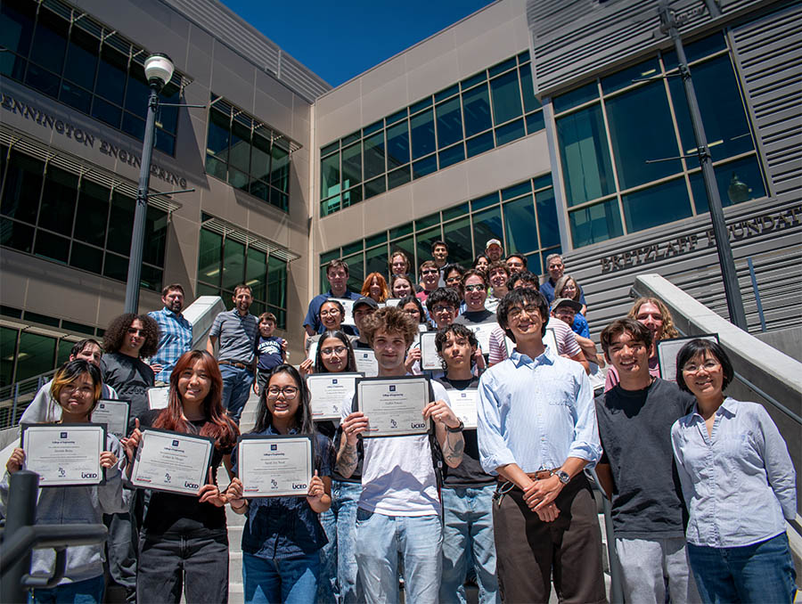 A group of about 30 people, some holding certificates, stand on an outdoor stairway, the William Pennington Engineering Building behind them.