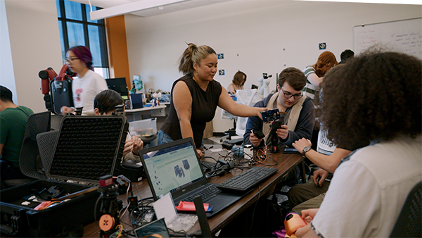 A woman stands in a robotics lab, providing assistance to seated students assembling small robots.