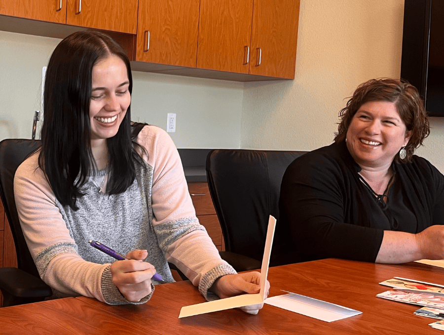 Two women are smiling at a table while signing cards.
