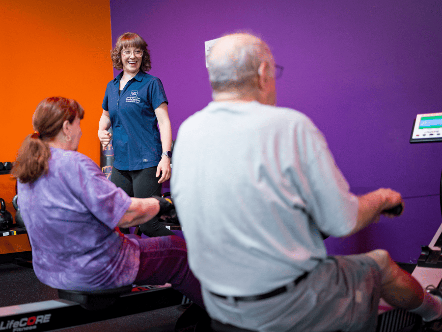 A woman stands in front of two older adults working out on rower machines.
