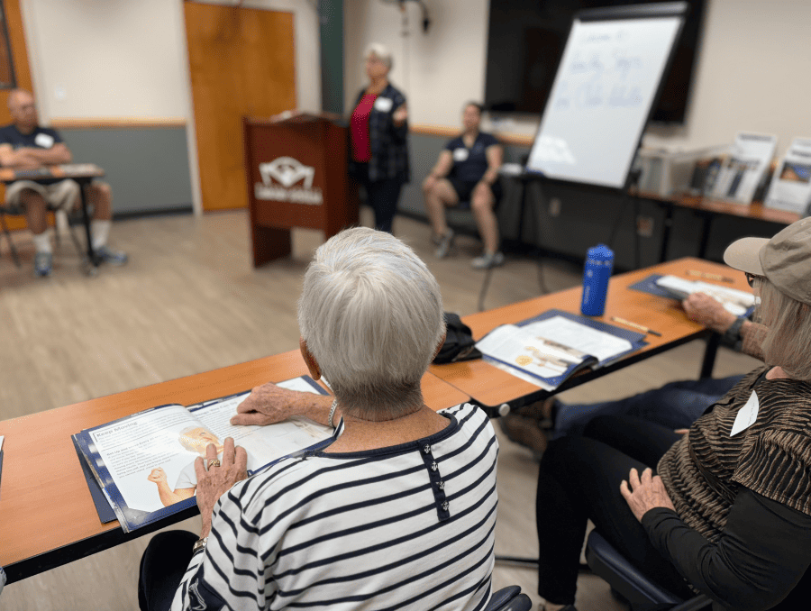 A group of older adults sit at a table looking at books and listening to a presentation in a classroom setting.