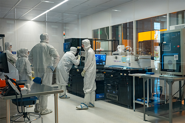 Five people in full protective gear stand in a cleanroom, one looking through a microscope.rough a microscope 