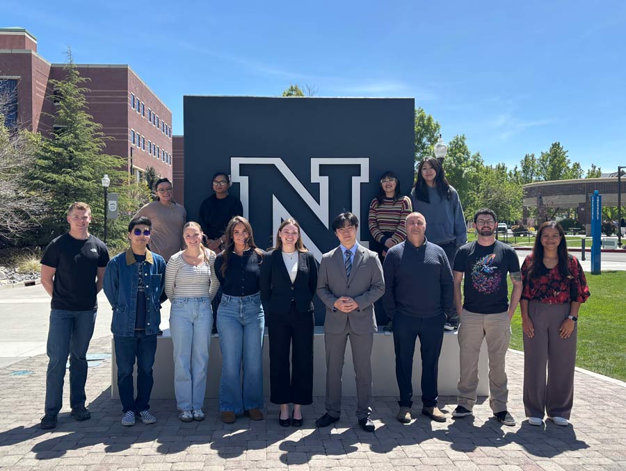 Thirteen people stand in front of and next to the Block N monument on the University campus on a sunny day, smiling for a photo.