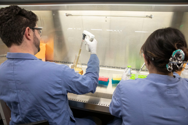 Two people sit with their backs to the camera, working in a fume hood. Both are wearing blue lab coats, protective eyewear and gloves and using pipettes.