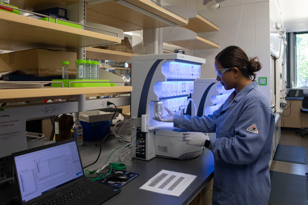 A researcher standing at the lab bench next to a laptop holding a bottle up to a machine. The researcher is wearing protective eyewear, gloves and a blue lab coat.