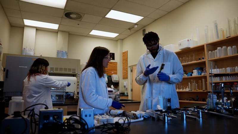 Presswood with two other researchers in lab coats in a laboratory looking at notes together.