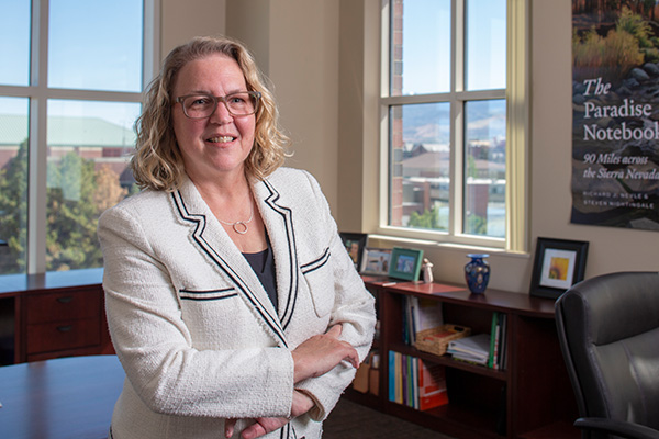 Dean Cardwell standing in her office in front of her desk. She is wearing a white blazer and she has her arms crossed in front of her body and she is smiling at the camera.