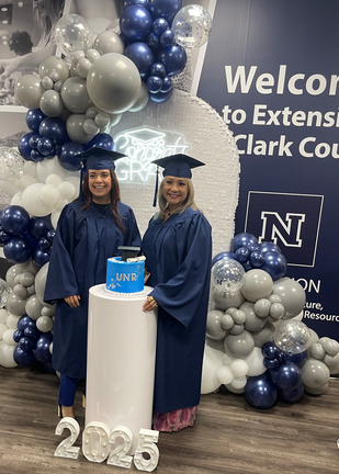 Two women in graduation gear posing amidst a background decorated with balloons.
