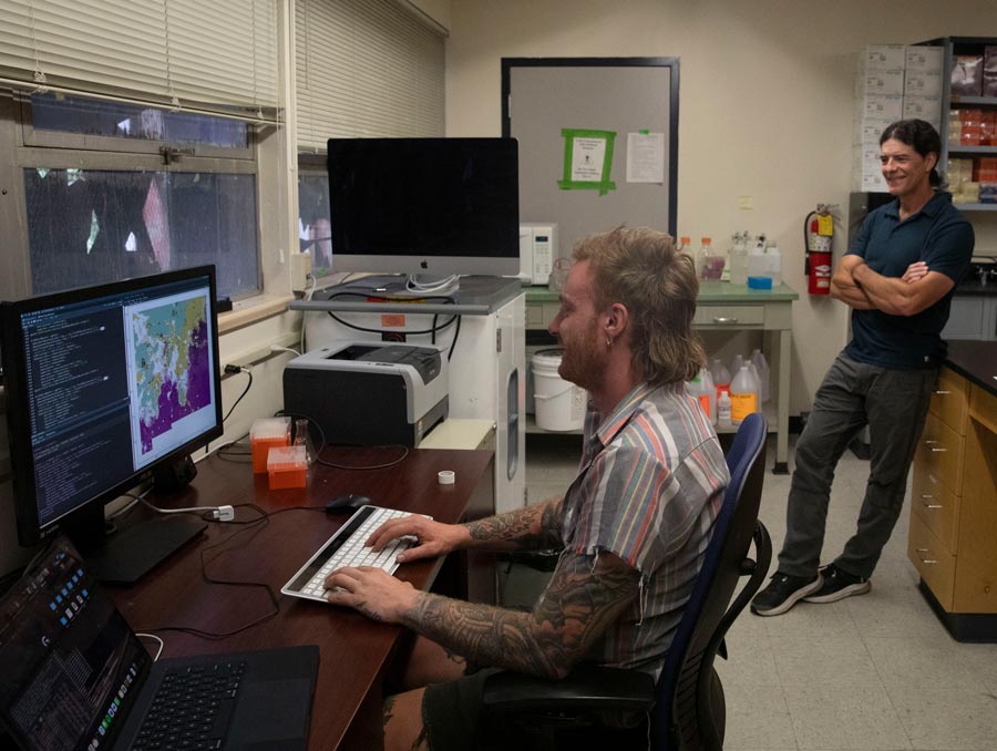 One person sitting at a computer in a research lab smiles while another person leans against a lab bench smiling with their arms crossed.