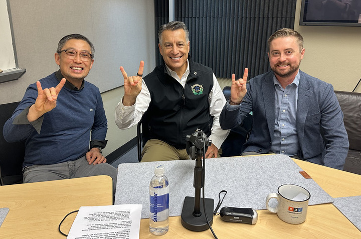Gi Yun sits next to President Sandoval and Brian Duggan in the podcast studio, all hold up Wolf Pack hand signs.