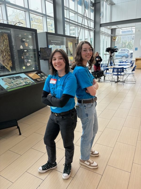 Two students in matching blue t-shirts smile toward the camera, back-to-back with their arms folded.