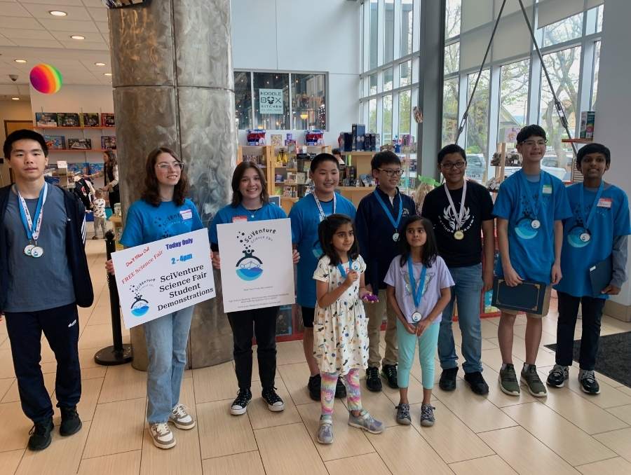 A group of K-12 students, some wearing matching blue t-shirts and some are wearing medals, smile for a photo. Two are holding signs with "SciVenture" on them.