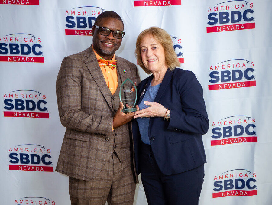 Shaundell Newsome and Nevada SBDC state director Winnie Dowling, smiling and holding an award in front of a white backdrop with the Nevada SBDC logo.