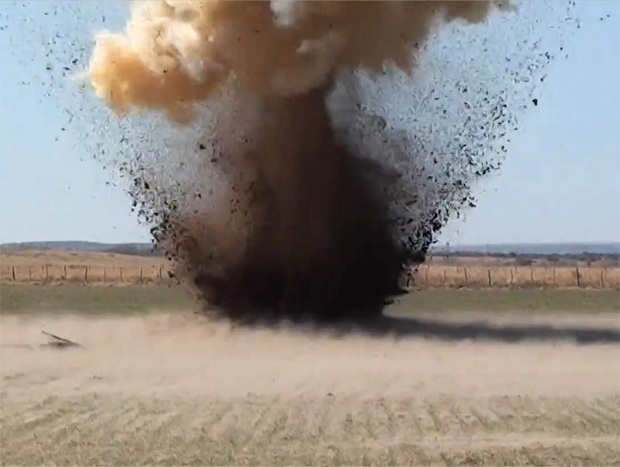 A large explosion in a desert setting, with dirt being thrown up from the ground and smoke on the top of the flying dirt.