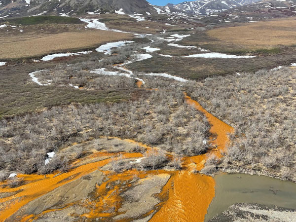 Water flowing at the base of a mountain with patches of snow is bright orange.