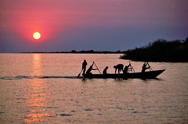 Silhouettes of six people rowing a boat to shore against a vibrant sunset.