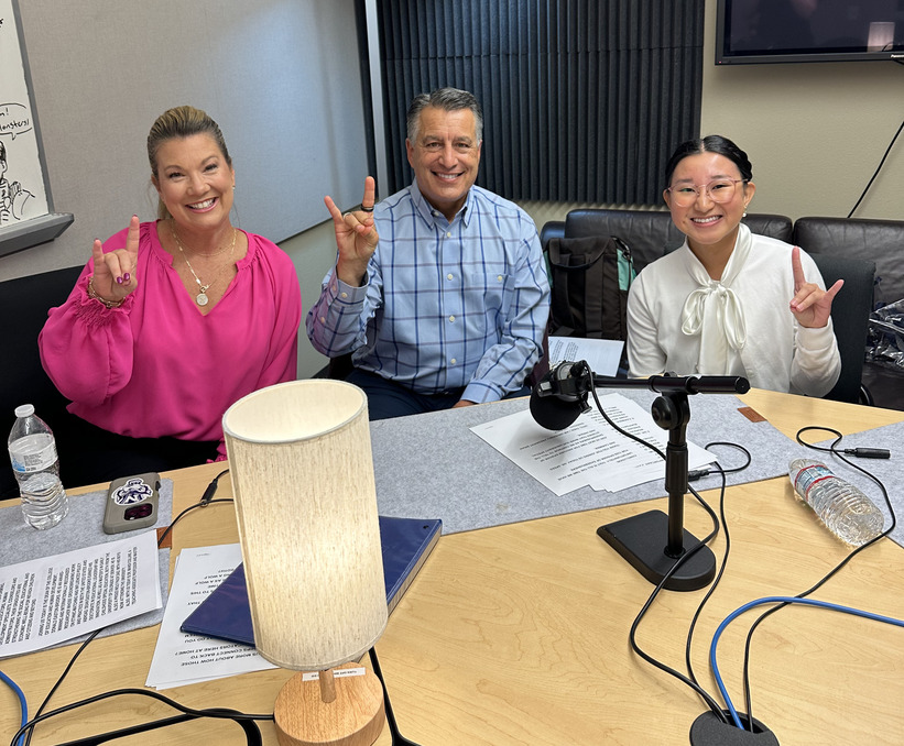 Heather Speed sits next to President Sandoval and Carmina Aglubat in the podcast studio, all hold up Wolf Pack hand signs.