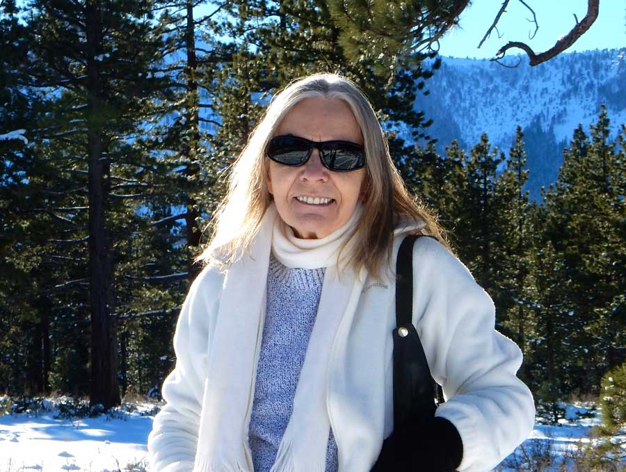 A photo of Jeanne Zeh smiling in a sunny, mountainous area with snow and trees behind her.