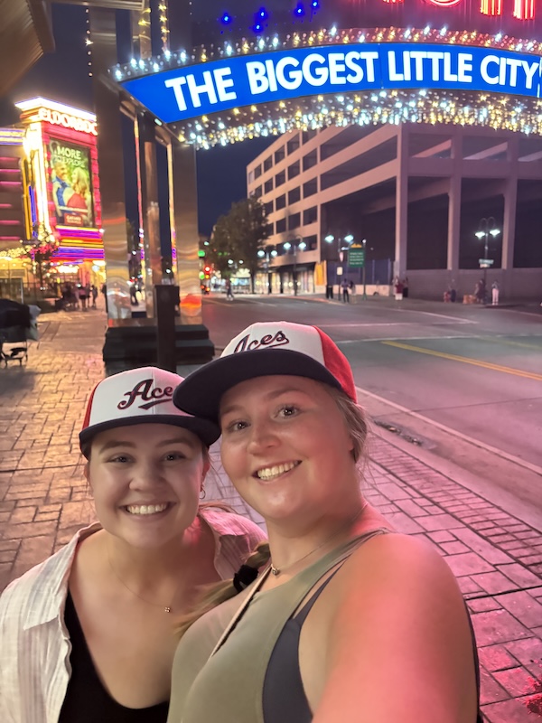 Mikayla and a friend wear Aces baseball hats and smile while standing under the Reno Arch sign at night.