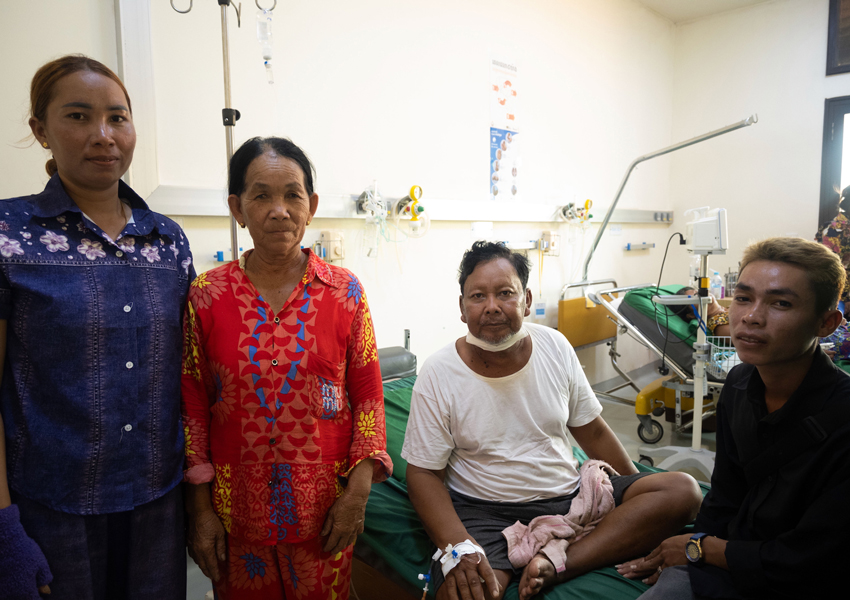 A family gathers around while one family member is seated in a hospital bed. 