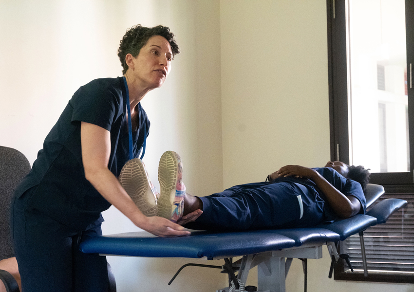 A nurse standing beside a bed lifting the leg of another nurse, demonstrating a part of nursing care. 