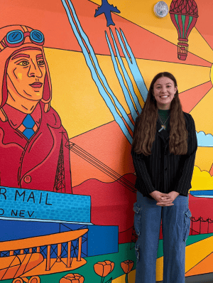 Reilly Moss standing in front of her mural that shows aviators with a sunrise behind them.
