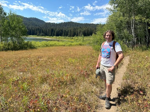 A person standing on a narrow dirt path through a grassy meadow near a lake, surrounded by green trees and forested hills. The sky is clear with scattered clouds. The person is wearing a white graphic T-shirt and holding a hat.