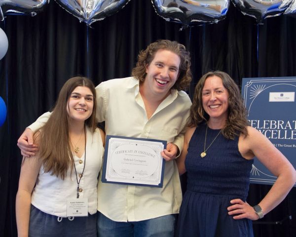 Three people stand together smiling at an event, with one person holding a certificate of achievement. They are surrounded by silver balloon decorations, and a sign in the background reads "Celebrate Excellence.